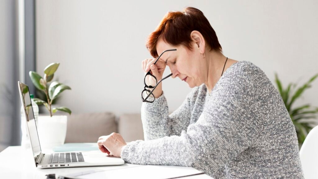 Una donna caucasica stanca con stress e cuore affaticato, 55 anni, con corti capelli castani, che tiene in mano degli occhiali davanti a un computer portatile in una stanza luminosa